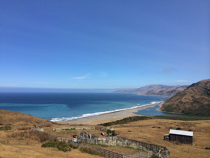 bright blue skies with wispy white clouds frame rolling tan hillsides with flat sections, an area of old rotting fences and an old shed lie in the foreground, a river meets the ocean in the distance